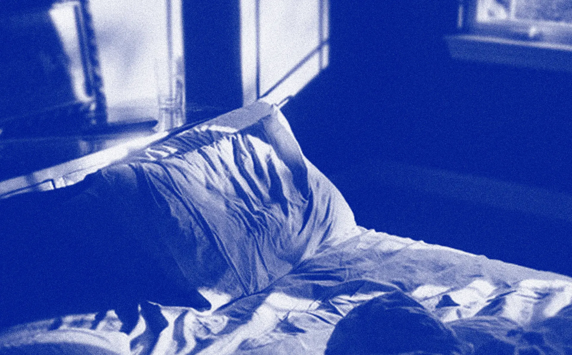 Blue-toned photo of a person asleep under rumpled bedding by a window