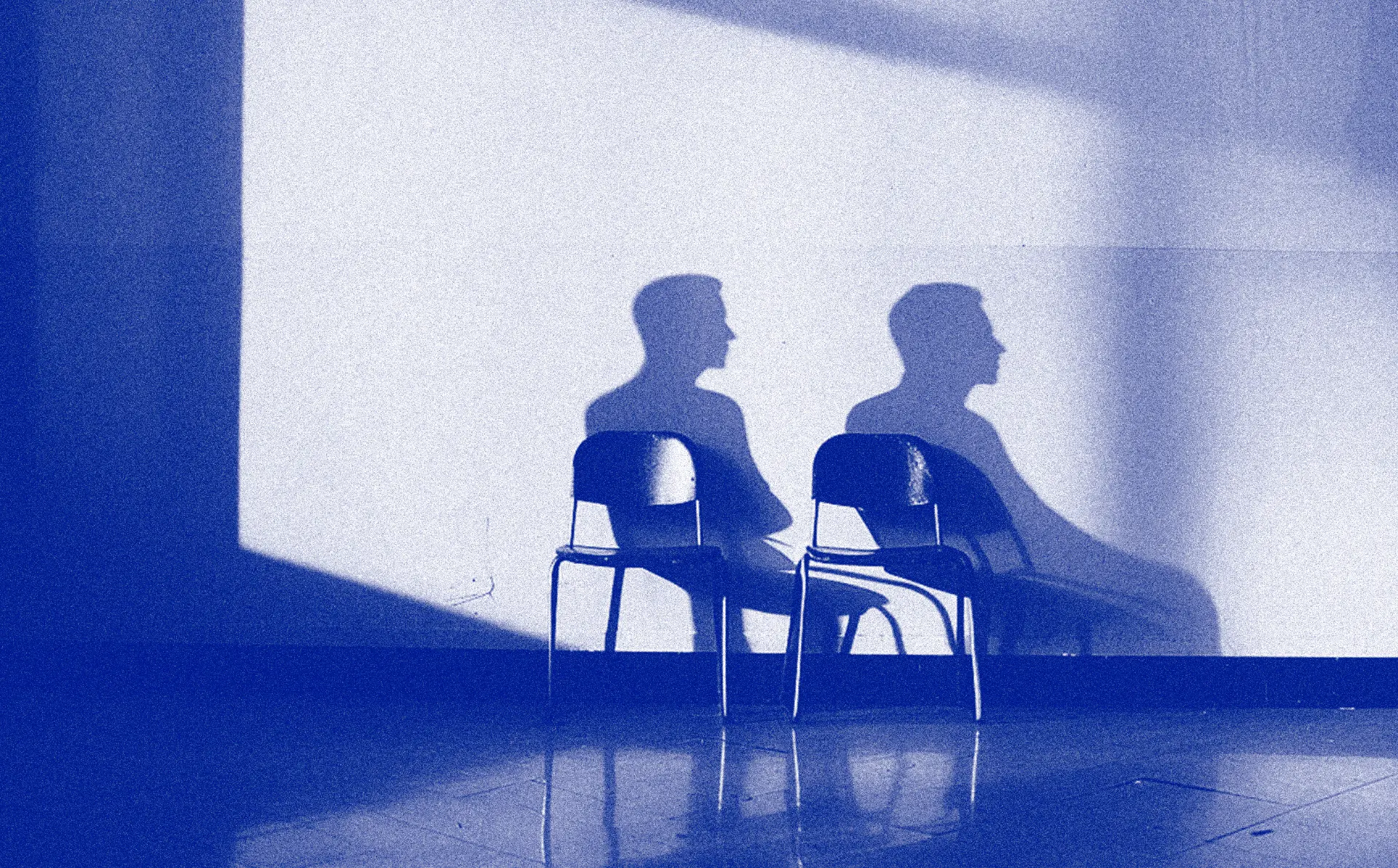 Two empty chairs against a wall, each with a person's shadow cast behind it.